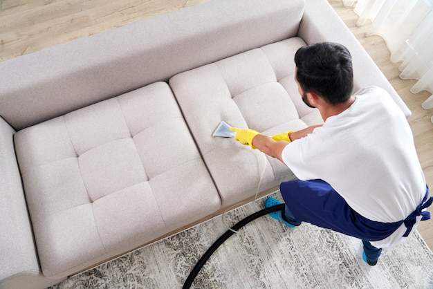 Photo close-up of housekeeper holding modern washing vacuum cleaner and cleaning dirty sofa with professionally detergent. professional springclean at home concept