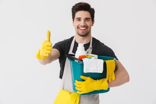 Photo handsome brunette houseman wearing apron standing isolated over white , holding bucket full of detergents