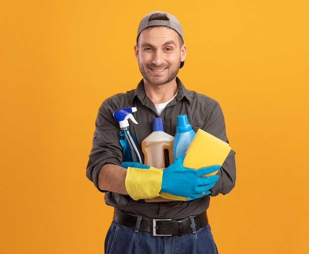 Free photo happy young cleaning man wearing casual clothes and cap in rubber gloves holding spray bottle and sponge  smiling cheerfully standing over orange wall