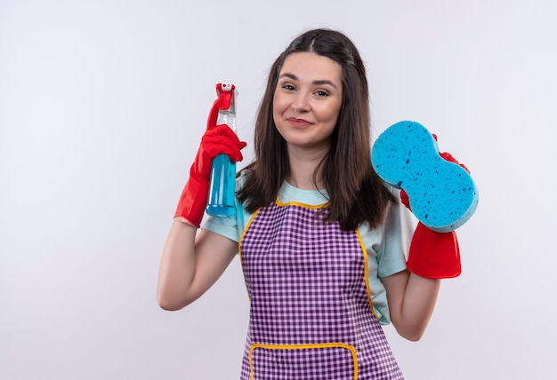Foto grátis linda jovem de avental e luvas de borracha segurando spray de limpeza e esponja olhando para a câmera com uma carinha feliz sorrindo