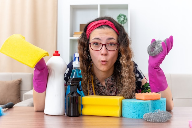Foto grátis mulher jovem com luvas de borracha, olhando para a câmera, confusa, sentada à mesa com materiais e ferramentas de limpeza na luz da sala de estar